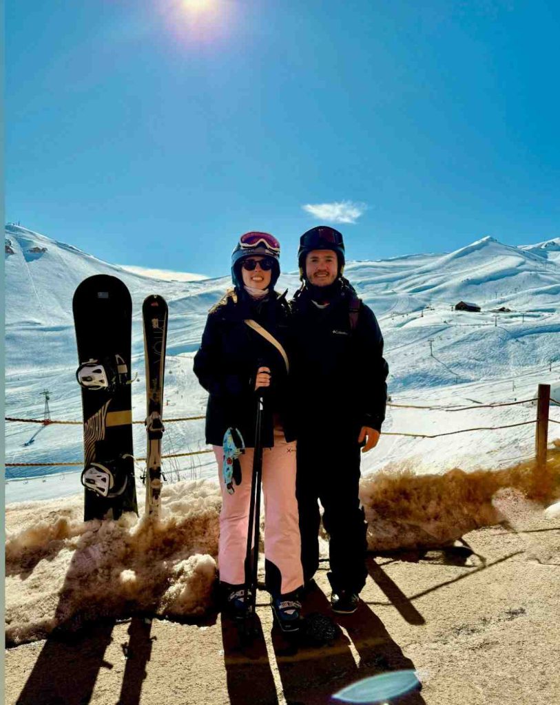 Pareja con equipos de esquí y snowboard en un centro de esquí de la cordillera, con cerros nevados de fondo